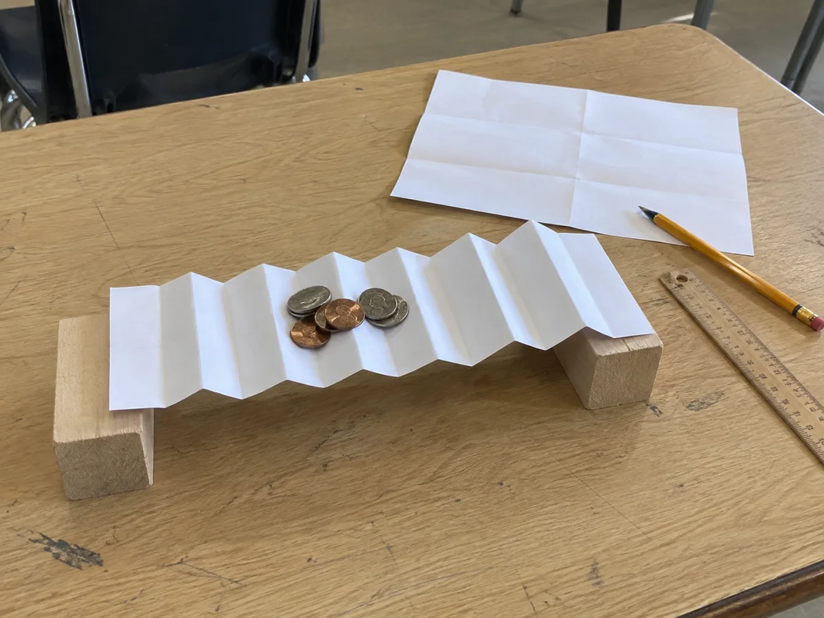 An accordion-folded paper bridge on two wooden blocks holds coins, with spare creased paper, a pencil and a ruler on a classroom desk.
